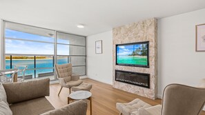 Ein modernes Wohnzimmer mit großem Fenster mit Blick auf einen Strand, einem Flachbildfernseher an einer Steinwand und einem Kamin.