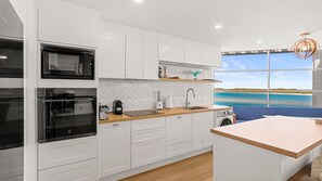 Eine moderne Küche mit weißen Schränken, einer Holz Arbeitsplatte und Blick auf einen Strand durch ein großes Fenster.