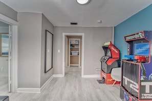 A room with a hallway leading to a door, a shelf with items, and a gaming console. - Historic Bungalow + Guest House • Patio & Grill (Tampa)