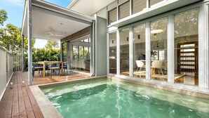 Zona de piscina exterior moderna amb un pont de fusta, portes de vidre que donen a un espai interior i un ventilador de sostre.