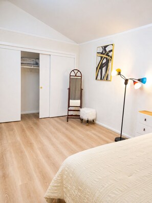 A bedroom with a bed, a wardrobe, a mirror, a stool, a floor lamp, and a painting on the wall.