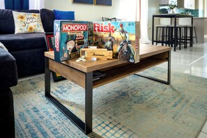 A living room with a coffee table holding board games Monopoly and Risk, a sofa with a floral pillow, and a kitchen in the background.