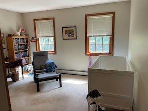 Ein Zimmer mit Bücherregal, einem Sessel, einem Babybett und zwei Fenstern mit Jalousien.