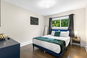Ein Schlafzimmer mit Bett, Nachttisch mit Lampe, Fenster mit Vorhängen und einem Wandbild.