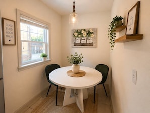 A dining area with a round table, two chairs, a hanging light fixture, a window with blinds, and wall-mounted shelves with plants and a framed sign.