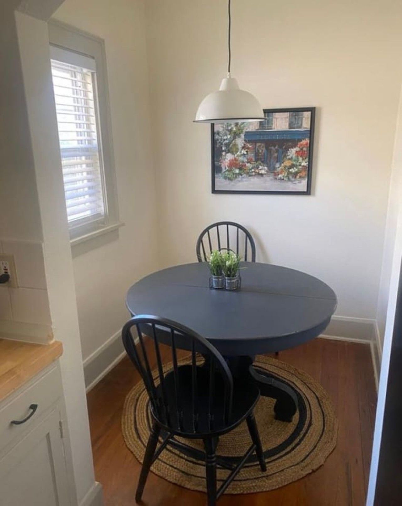 Un coin repas avec une table ronde, deux chaises, un luminaire suspendu, un tableau encadré au mur et une fenêtre avec des stores.