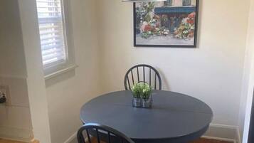 A dining area with a round table, two chairs, a hanging light fixture, a framed picture on the wall, and a window with blinds.