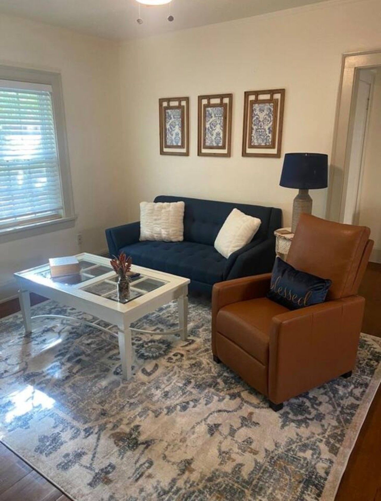 Un salon avec un canapé bleu foncé, un fauteuil en cuir marron, une table basse blanche et un tapis à motifs. Trois tableaux encadrés sont accrochés au mur au-dessus du canapé.