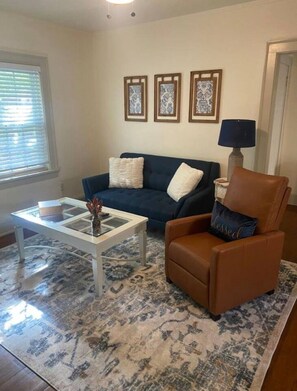 A living room with a dark blue sofa, a brown leather chair, a white coffee table, and a patterned rug. There are three framed pictures on the wall above the sofa.