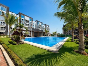 A swimming pool surrounded by well-maintained grass and palm trees, with modern buildings in the background.