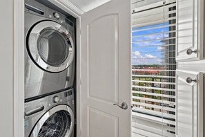 A laundry room with two front-loading washing machines, a door, and a view through a window with blinds. - Edgewater T2 703 - 3 Bedrooms! (Panama City Beach)