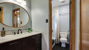 A bathroom with a marble sink, dark wooden cabinets, a round mirror, and a visible shower area. - Theater, Spa, Gym, & Vinyls: 2 Homes, 1 Epic Stay! (Austin)