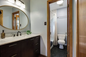 A bathroom with a marble sink, dark wooden cabinets, a round mirror, and a visible shower area. - Theater, Spa, Gym, & Vinyls: 2 Homes, 1 Epic Stay! (Austin)