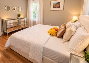 Ein Schlafzimmer mit einem großen Bett, einer Kommode, einem Fenster mit Vorhängen und einem gerahmten Bild an der Wand.