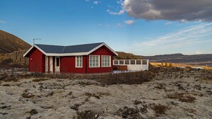 Una cabaña roja con un porche blanco, rodeada de terreno rocoso y montañas.