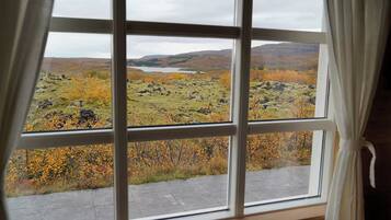 Una ventana con vistas al paisaje, cortinas entreabiertas y un camino de piedra que conduce al exterior.
