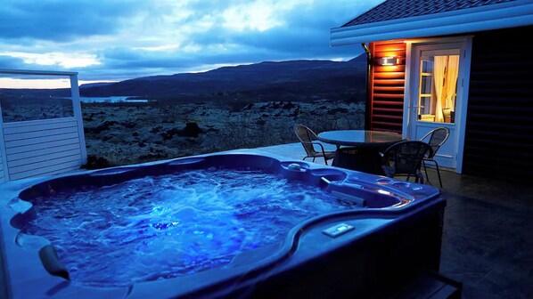 A hot tub with a view of a mountainous landscape, a table with chairs, and a door with a light on.