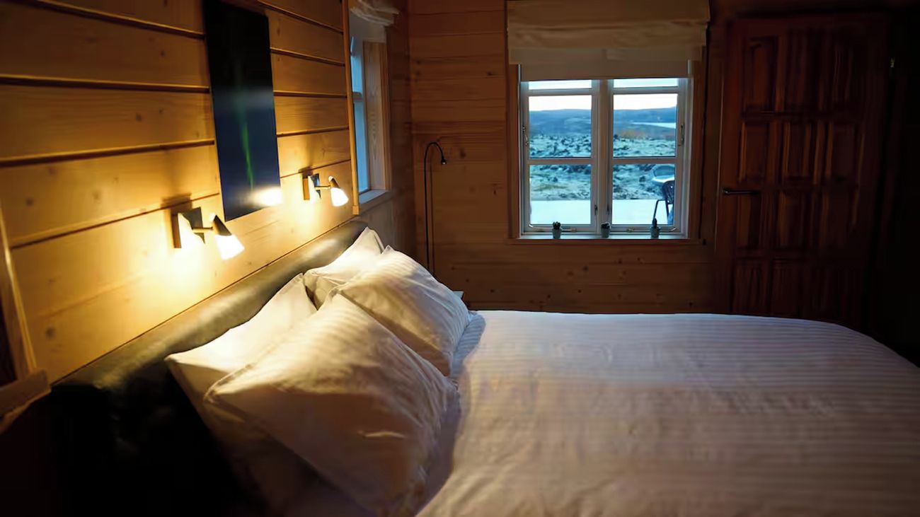 A wooden cabin bedroom with a bed, a window with a view, and wall-mounted lights.