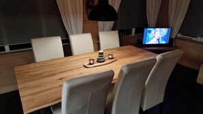 A dining area with a wooden table, white chairs, a black pendant lamp, and a television displaying a person.