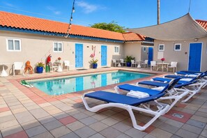 A swimming pool with lounge chairs, a shaded area, and a building with blue doors. - Aruba Oasis w/ Pool · King Bed · #14 (Aruba)