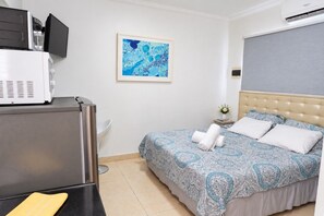 A hotel room with a bed, a desk, a chair, a small table with a vase, and a wall-mounted air conditioner. - Aruba Oasis w/ Pool · King Bed · #14 (Aruba)