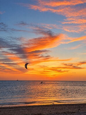 Une scène de plage au coucher du soleil, avec une personne sur une planche à pagaie et un voilier à l’horizon.