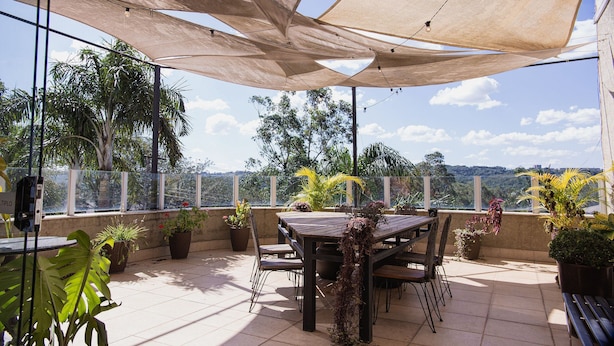 Un patio al aire libre con una mesa y sillas de madera, rodeado de plantas en macetas, bajo grandes toldos de sombra.