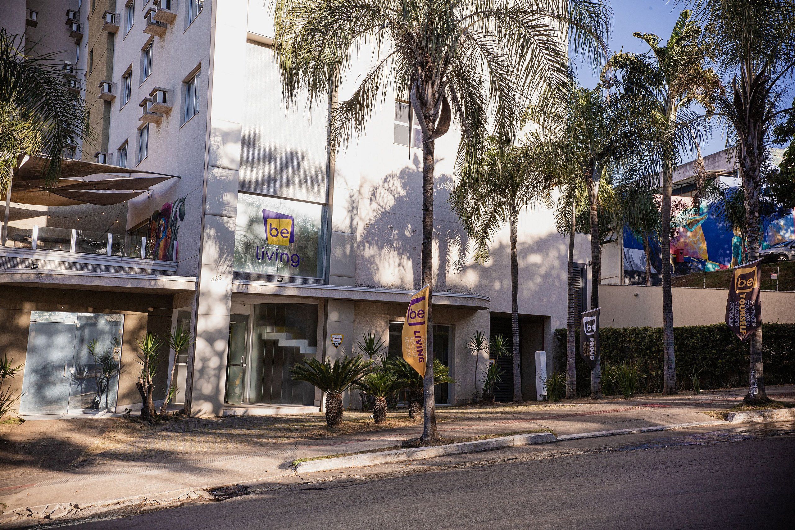 Um edifício moderno com fachada de vidro extensa, palmeiras e uma placa com a inscrição 'be living'.