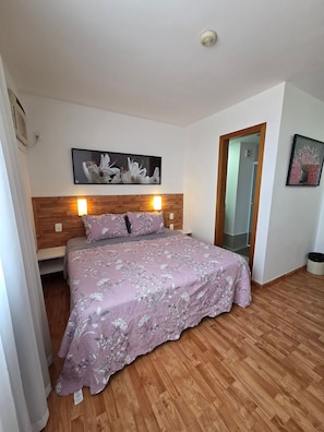 Un dormitorio con cabecera de madera, cama con ropa de cama floral, un cuadro colgado en la pared y una lámpara de techo.