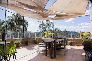Un patio al aire libre con una mesa y sillas de madera, rodeado de plantas en macetas, bajo grandes toldos de sombra.