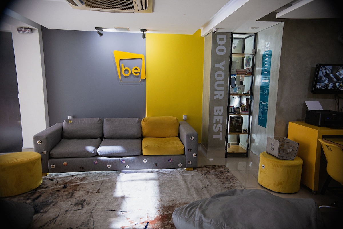 Una sala de estar moderna con un sofá gris, cojines amarillos y una pared grande con el logo 'be'. Hay una estantería con varios objetos y un televisor sobre un mueble.