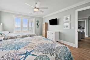 A bedroom with a large bed, a ceiling fan, a TV, and a door leading to a bathroom. - Modern Oceanfront 5BR Home, Private Pool, Elevator, Sleeps 18, Ocean Drive Location (North Myrtle Beach)