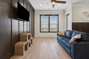A modern living room with a flat-screen TV, a ceiling fan, and a large window with blinds. - Tides & Vibes: Spacious 5BR Retreat w/ Private Pool, across the street from the beach (North Myrtle Beach)