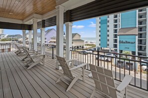 Une terrasse extérieure couverte, avec des chaises en bois, donnant sur une plage, et un bâtiment portant l’inscription « Shalimar 2908 » en arrière-plan.