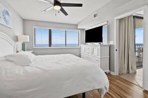 Ein Schlafzimmer mit einem Bett, einem Deckenventilator, einem Fernseher, einer Kommode und einem Fenster mit Jalousien.