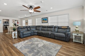 A modern living room with a sectional sofa, a ceiling fan, a dining area with a table and chairs, and a kitchen in the background. - 6BR ocean-view home in Crescent Beach with beach access and parking (North Myrtle Beach)