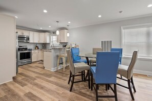 A modern kitchen with built-in appliances, a dining area with blue chairs, and a large window with blinds. - 6BR ocean-view home in Crescent Beach with beach access and parking (North Myrtle Beach)