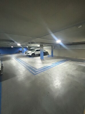 An underground parking garage with concrete floors, blue painted lines, and a white car parked in the center. - Melhores Flats - Acolhedor, 30m do mar  (João Pessoa)