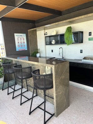 A modern kitchen with a marble countertop, a sink, and a bar area with stools. - Melhores Flats - Acolhedor, 30m do mar  (João Pessoa)