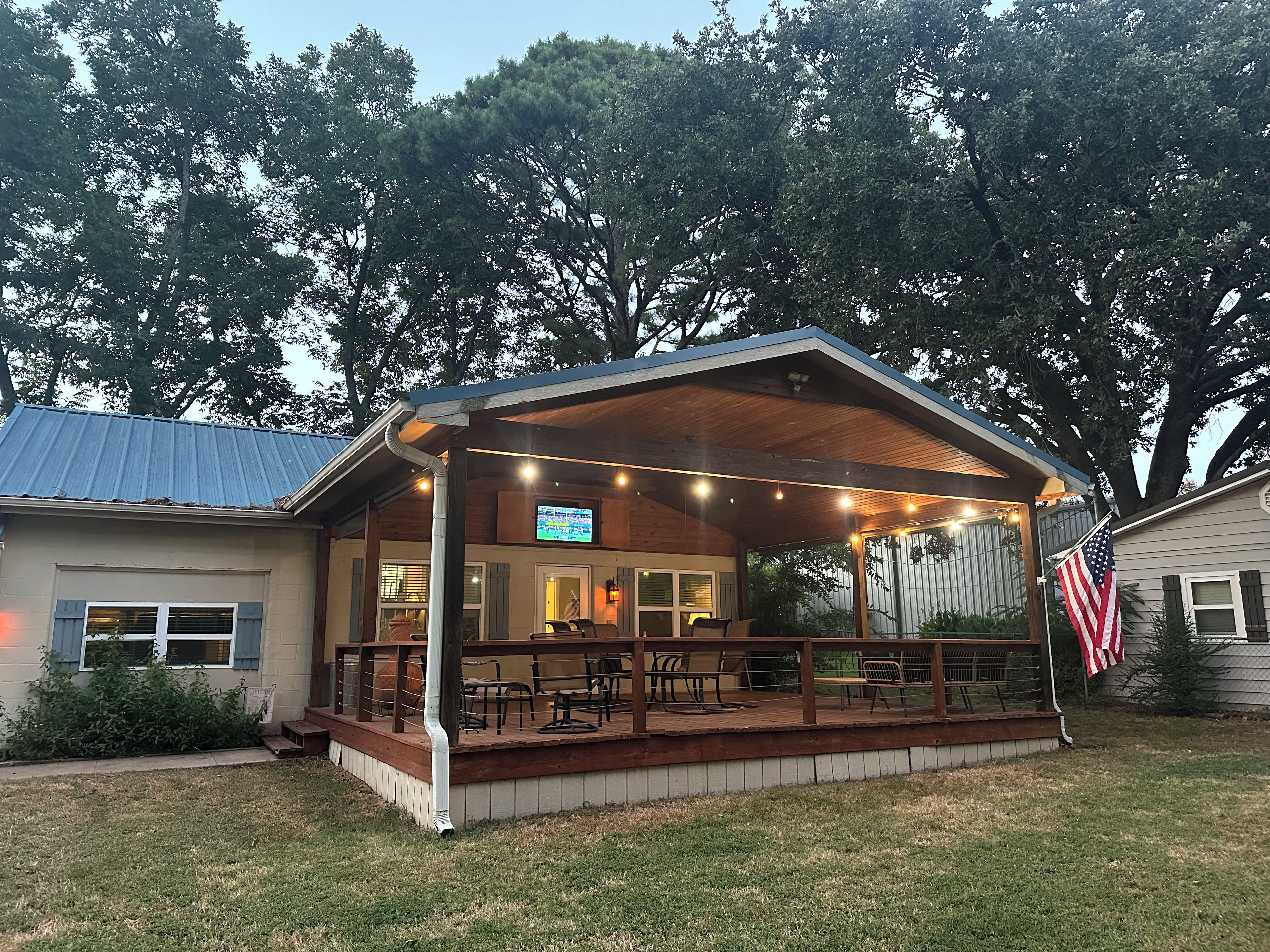 A covered outdoor seating area with tables and chairs, an American flag, and a television screen.