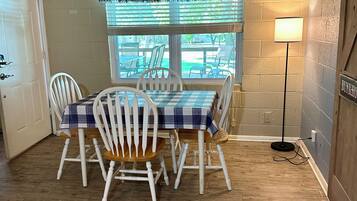 Un comedor con una mesa dispuesta para cuatro, sillas blancas, un ventanal con persianas y una lámpara de pie.