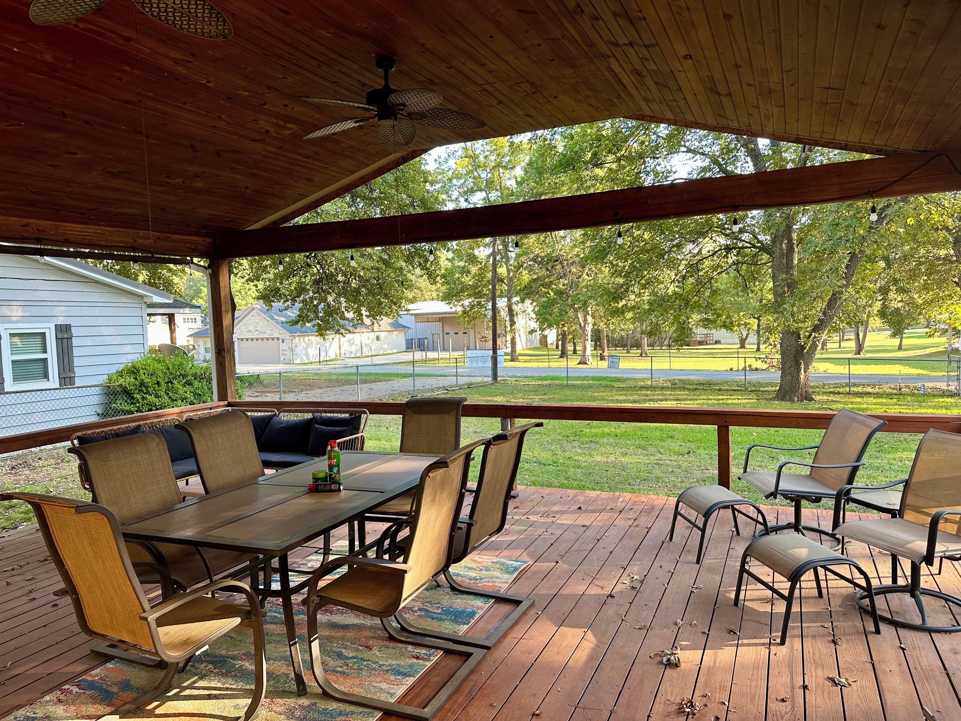 Un patio techado al aire libre con una mesa y sillas, un ventilador de techo y vistas a una zona residencial con árboles y casas.
