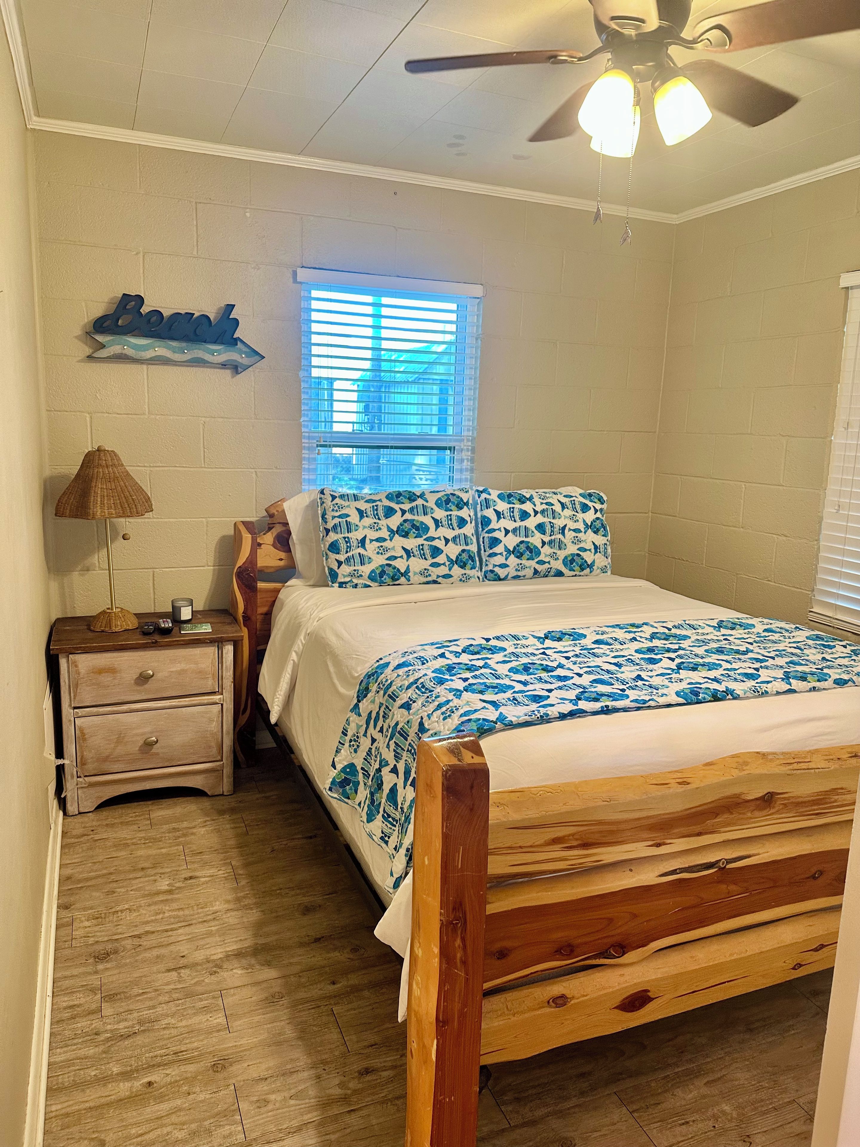 Un dormitorio con una cama de madera, una mesita de noche, una lámpara, un ventilador de techo y una ventana con persianas.