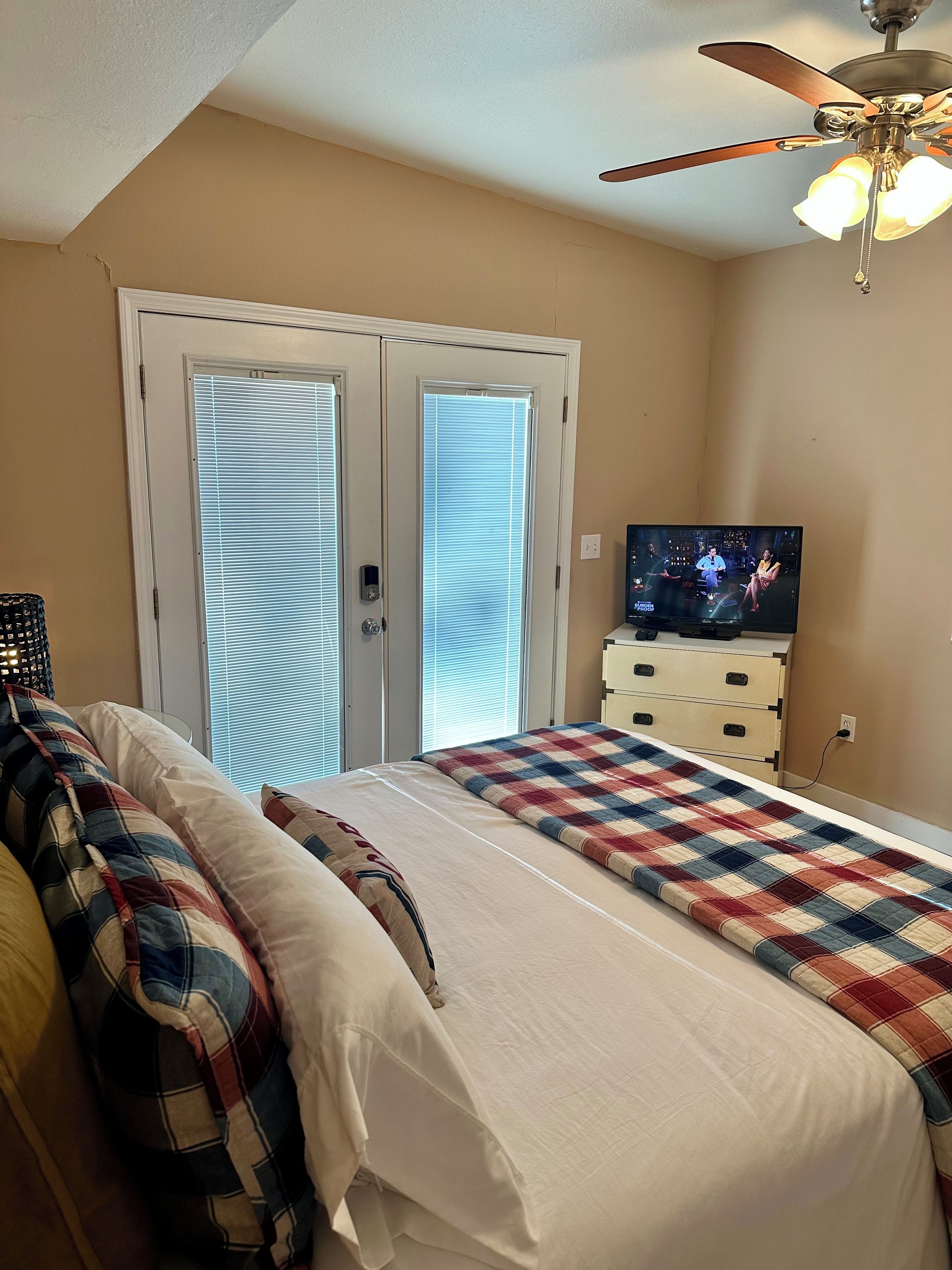 A bedroom with a bed, a television, a ceiling fan, and a door with blinds.
