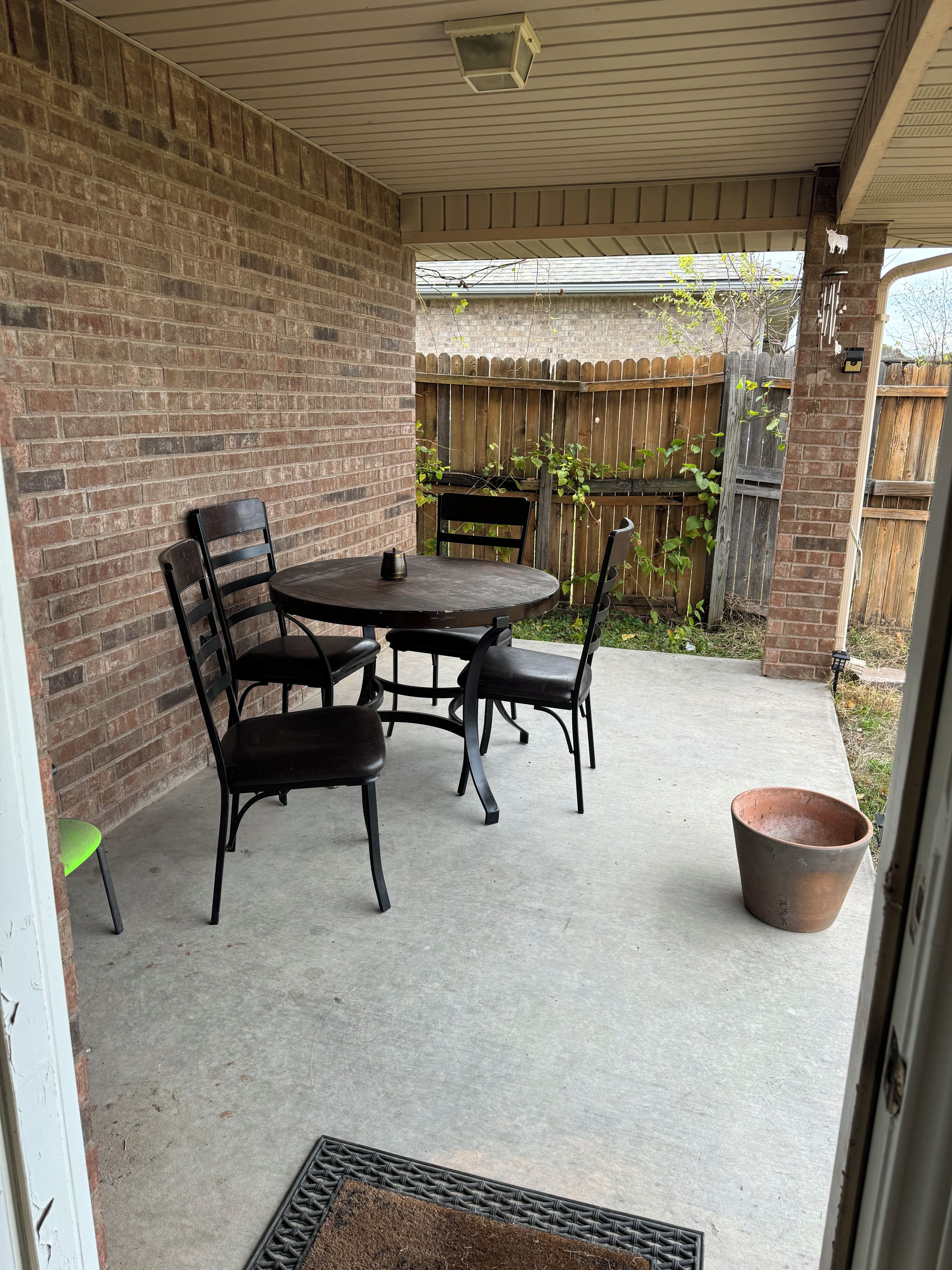 Een overdekt terras met een bakstenen muur, een houten schutting en een tafel met stoelen.