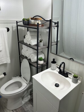 A bathroom with a white sink, a toilet, a black shelf unit with towels and plants, and a mirror. - Hillside Hideaway 3 Bed 3 Bath home stocked with all the comforts of home (Natchez)