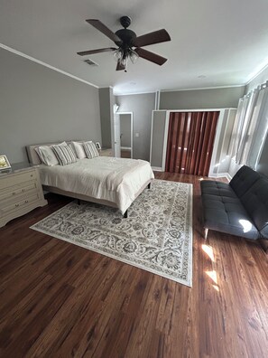 A bedroom with a bed, a ceiling fan, a dark sofa, a patterned rug, and wooden flooring. - Hillside Hideaway 3 Bed 3 Bath home stocked with all the comforts of home (Natchez)
