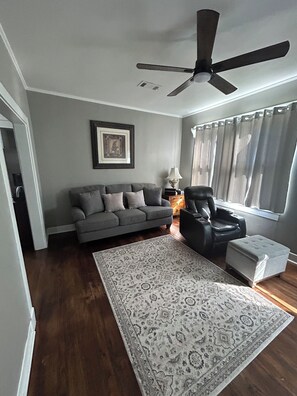 A living room with a grey sofa, a black leather chair, a patterned rug, a ceiling fan, and a window with curtains. - Hillside Hideaway 3 Bed 3 Bath home stocked with all the comforts of home (Natchez)