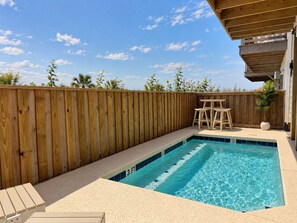 Ein Schwimmbecken mit Holzterrasse und Liegestühlen, umgeben von einem Holzzaun und blauem Himmel.