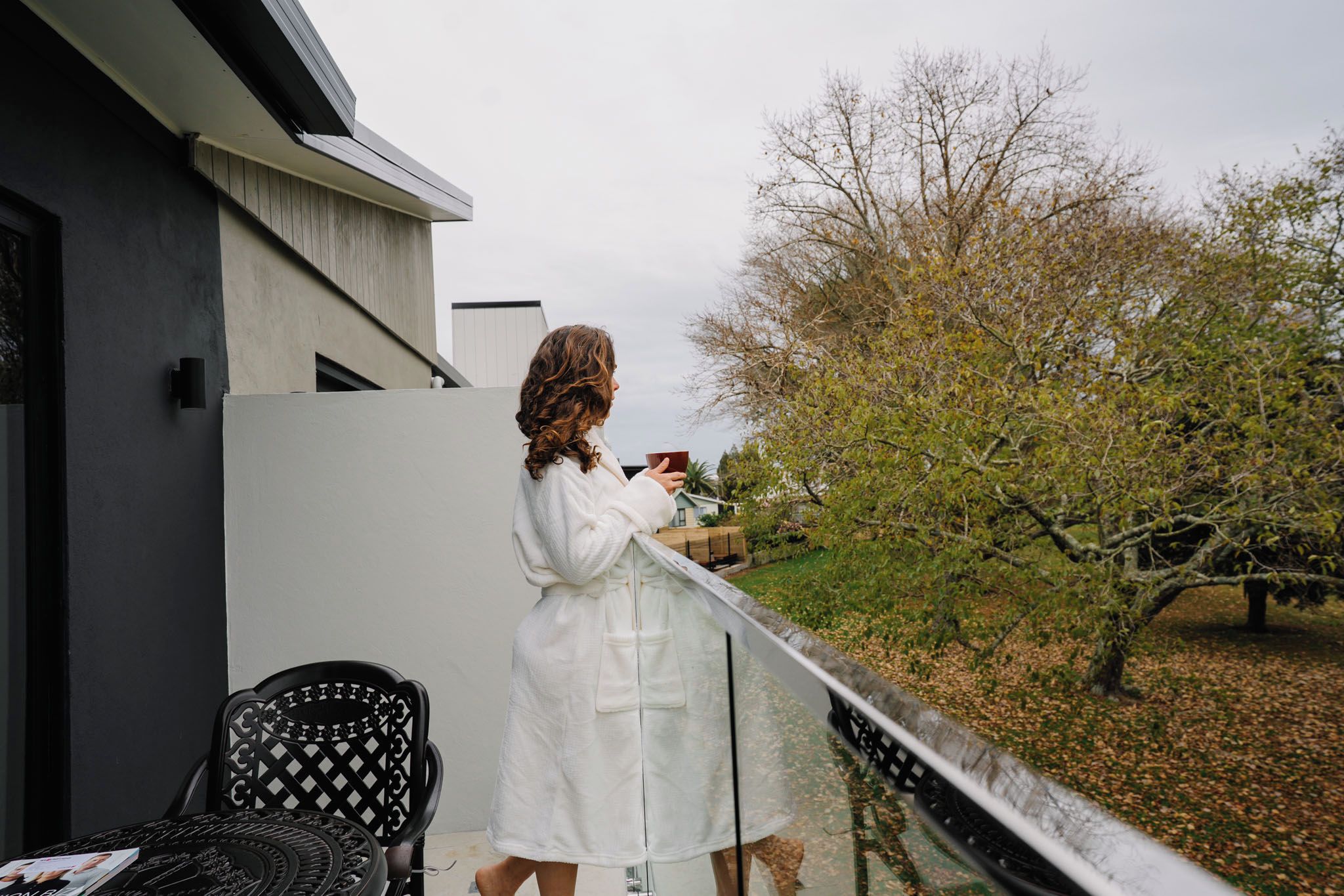 A person standing on a balcony with a railing, holding a cup, with a building and trees in the background.